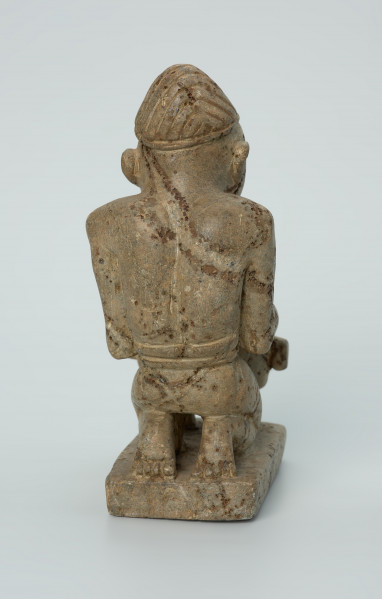figura kultu przodków - Ujęcie z tyłu. Rzeźbiona w szarym kamieniu postać mężczyzny w pozycji klęczącej, grającego na bębnie szczelinowym tam-tamie. Widoczne rysy, mikropęknięcia, liczne malutkie otworki, przebarwienia, ślady klejenia oraz ubytek na lewym łokciu.