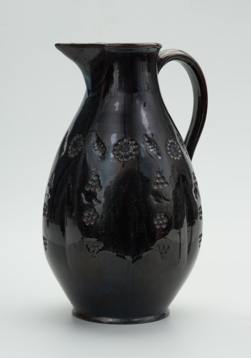 gliniany dzban zdobiony motywem roślinnym - Ujęcie z lewej strony. Dzban wykonany z gliny. Brzusiec łagodną linią przechodzi w szyjkę o pogrubionej krawędzi wylewu, z której wyodrębniony jest duży, ostro zakończony dziobek. Ucho taśmowate, wzdłużne, mocowane w połowie wysokości szyjki i w połowie wysokości brzuśca. Brzusiec zdobiony wykonanym techniką stempelkową ornamentem o motywie roślinnym. Wzór ujęty w trzy poziome pasy: dolny składa się ze schematycznie przedstawionych gron, środkowy - z pionowych elementów, na które składają się: grona, gałązka i pięciopłatkowy kwiatek, górny - z ułożonych naprzemiennie wielopłatkowych kwiatów i ulistnionych gałązek. Przy krawędzi szyjka zdobiona drobnym wytłaczanym szlaczkiem. Całość szkliwiona.