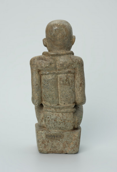 Ujęcie z tyłu. Rzeźbiona w biało-szarym kamieniu dość duża postać kobiety w pozycji siedzącej z nogami skrzyżowanymi. Kobieta siedzi na małej, kwadratowej, lekko zaokrąglonej na rogach podstawie, w dłoniach trzyma dziecko. Widoczne rysy, mikropęknięcia i liczne malutkie otworki.