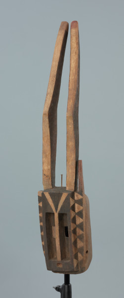 maska - Ujęcie z przodu, z lewej strony. Drewniana, rzeźbiona maska antylopy, z dużymi geometrycznymi otworami na oczy.