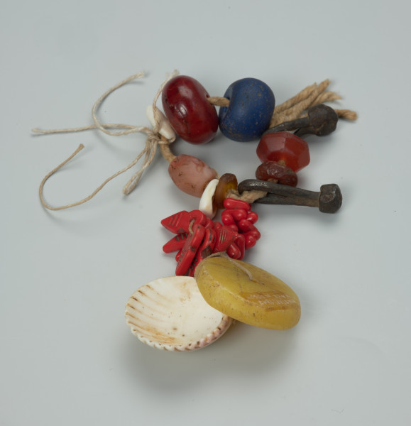 biżuteria, bransoleta - Ujęcie z przodu. Zamknięta, kolorowa bransoleta z wisiorkiem. Na bawełnianym sznurku znajduje się wiele różnych elementów: żelazne ogniwa, szklane i kamienne paciorki o różnych rozmiarach i kolorach, muszelki kauri. Do bransolety dołączono wisior składający się z kilkunastu elementów naniesionych na bawełnianą nić: czerwonych paciorków o różnych kształtach, dużego, okrągłego, płaskiego i żółtego paciorka oraz większej muszli.