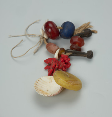 biżuteria, bransoleta - Ujęcie z przodu. Zamknięta, kolorowa bransoleta z wisiorkiem. Na bawełnianym sznurku znajduje się wiele różnych elementów: żelazne ogniwa, szklane i kamienne paciorki o różnych rozmiarach i kolorach, muszelki kauri. Do bransolety dołączono wisior składający się z kilkunastu elementów naniesionych na bawełnianą nić: czerwonych paciorków o różnych kształtach, dużego, okrągłego, płaskiego i żółtego paciorka oraz większej muszli.