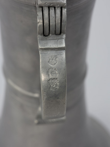 Cynowy dzban o kształcie cylindrycznym - detal; Ucho taśmowe, silnie esowato wygięte, zakończone wałkiem. W górnej części ucha zawias podtrzymujący pokrywę z uchwytem wygiętym esowato i ozdobioną płaskorzeźbą przedstawiającą lwią głowę.