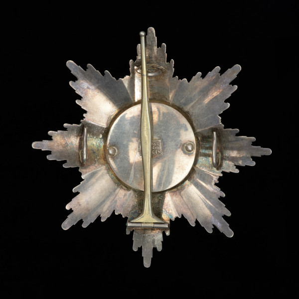 Gwiazda Orderu Grobu Świętego - Ujęcie z tyłu. Ośmiopromienna gwiazda o falistych promieniach z krzyżem jerozolimskim w środku. Na odwrocie szpila do zapinania, uchwyty do szarfy oraz sygnatura firmy jubilerskiej Godeta w Berlinie.