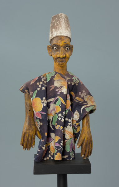 lalka teatralna: Kalhaldi (muzułmański uczony) - Ujęcie z przodu. Lalka teatralna przedstawiająca postać męską w nakryciu głowy w formie walca rozszerzonego u dołu, pomalowanego na biało. Szata z kwiecistego materiału, szeroka, z szerokimi rękawami. Oczy białe, z czarnymi źrenicami, drobne i wypukłe, z zaznaczonymi powiekami dolnymi oraz łukowatymi czarnymi brwiami.  Nos rozszerzony u dołu, usta zamknięte, z dolną wargą pełniejszą. Ręce przy tułowiu. Dłonie z zaznaczonymi palcami.