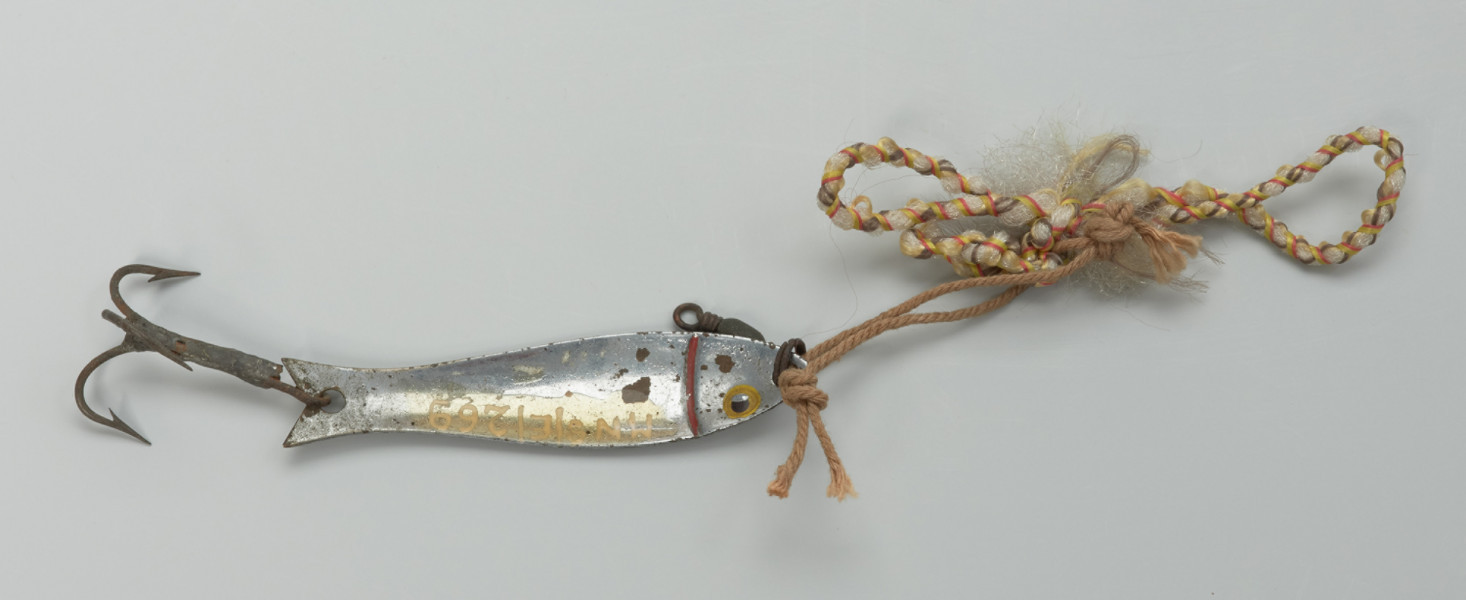 sztuczna przynęta (błyskawka) - Ujęcie tyłu z góry. Sztuczna przynęta w kształcie rybki wycięta z cienkiej, cynowej blaszki. Lekko wypukła. W przedniej i tylnej części znajdują się okrągłe otwory, do których mocowane są: w przedniej – kółeczko na żyłkę, w tylnej – haczyk w postaci wykonanej z metalu trójramiennej kotwiczki z zadziorami.