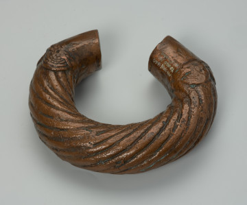 biżuteria, bransoleta - Ujęcie z tyłu góry. Otwarta, miedziana, owalna bransoleta ozdobiona grubymi, zaokrąglonymi, biegnącymi dookolnie prążkami; cienkimi, ukośnymi nacięciami oraz półkolami przypominającymi wachlarze wyłaniające się z motywu plecionki.