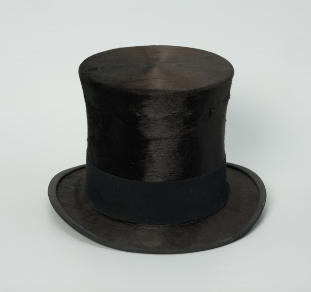 czarny cylinder z welurowej tkaniny - Ujęcie z przodu; Czarny cylinder z welurowej tkaniny z czarnym otokiem ponad rondem wykończonym pasmanterią. Wysoki cylinder, sztywny, z główką w kształcie walca oraz małym rondem.