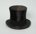 czarny cylinder z welurowej tkaniny - Ujęcie z przodu; Czarny cylinder z welurowej tkaniny z czarnym otokiem ponad rondem wykończonym pasmanterią. Wysoki cylinder, sztywny, z główką w kształcie walca oraz małym rondem.