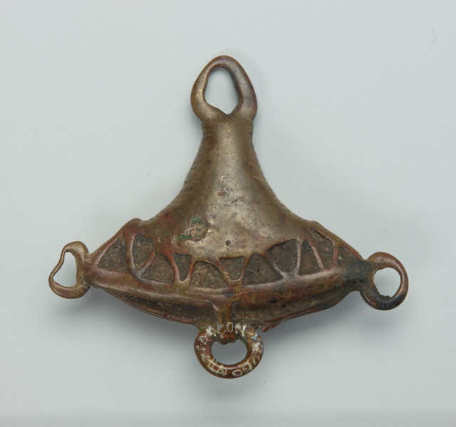 biżuteria, ozdoba - Ujęcie z przodu. Mały, trójkątny, ozdobiony wzorami geometrycznymi wisiorek, wykonany z brązu. W jego dolnej części udekorowany został trzema małymi, pustymi wewnątrz kółeczkami (jedno lekko zdeformowane). W jego górnej części wykonano owalny otwór. Widoczne ślady użytkowania, lekko wytarty wzór.