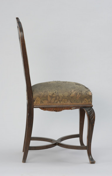 krzesło - Ujęcie z lewego boku. Krzesło w stylu neobarokowym, oparcie i siedzisko tapicerowane. Nogi zwężające się do dołu, przednie wygięte kabriolowo, górne wybrzuszenie ozdobione płaskorzeźbionym motywem palmety, zakończenie dolne trójkątne. Tylne nogi lekko wygięte, powiązane są z przednimi krzyżową poprzeczką umieszczoną u dołu, składającą się z dwóch stykających się w środku półokrągłych pałąkowych listew. Konstrukcja zaplecka stanowi pionowe przedłużenie tylnych nóg o profilowanych krawędziach, zamykających się dwoma półkołami w kształt serca, ponad którym wyprowadzono łukowe zwieńczenie w formie mięsiście rzeźbionego, rozłożonego na boki bukietu liści. Poniżej sercowego styku listew bocznych wydłużony w pionie medalion opasany płaską listwą z wypełnieniem tapicerką. Tkanina w kwiatowy wzór w odcieniach zieleni, różu, żółcieni i ciemnego popielu. Pomiędzy owalem medalionu i listwami oparcia ażurowe wypełnienie mięsistą płaskorzeźbą o motywach roślinnych. Taką samą tkaniną obite jest siedzisko o konstrukcji sprężynowej, stosunkowo wysokie, poszerzające się do przodu, w dolnej części wykończone półwałkiem i pasmanterią, kryjącą krawędź tapicerki.