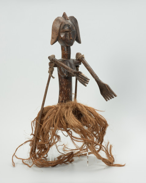 Ujęcie z przodu z prawej strony. Lalka umieszczona na koszyku-czapeczce pokrytej włóknami roślinnymi, noszona na głowie, przedstawiająca kobietę od pasa w górę. Linia brwi i nos wyodrębnione z twarzy. Usta lekko zaznaczone. Twarz ozdobiona skaryfikacją: po dwie linie pionowe na czole i policzkach. Ręce ruchome, poruszają się w linii poziomej za pomocą patyków umocowanych w 