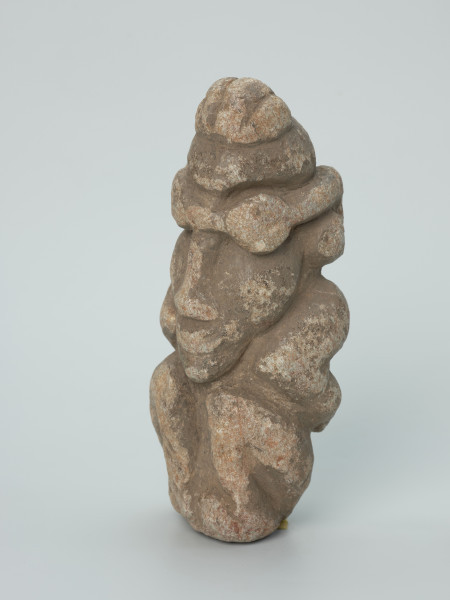 rzeźba; Figura kultu zmarłych - Ujęcie z przodu z lewej strony. Rzeźbiona w szarobeżowym steatycie siedząca postać ludzka z dużą głową o masywnych, okragłych okularach i przykurczonymi nogami, których kolana sięgają brody.