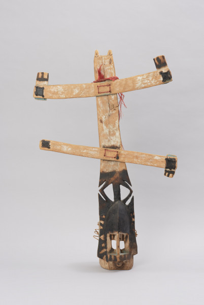 maska kanaga - kompletna - Ujęcie z przodu. Maska wykonana z materiałów organicznych: skóry, włókna roślinnego i drewna. Pionowa część w górnej połowie wypełniona białymi znakami, dolna część pomalowana na czarno. Do części pionowej przymocowane dwie listwy poziome również pomalowane w białe znaki.