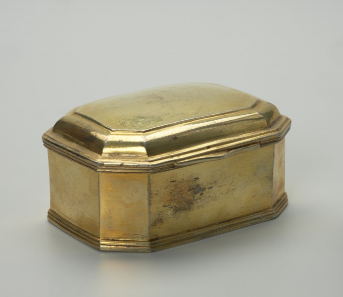 pozłacane puzderko na niskim, profilowanym cokoliku - Ujęcie z przodu, z ukosa; Pozłacane puzderko na niskim, profilowanym cokoliku. Korpus ośmioboczny, na rzucie prostokąta ze ściętymi narożami. Pokrywa na zawiasie, z dwoma uskokami, lekko wybrzuszona z wielokrotnie profilowanym brzegiem, z przodu w centralnej części wysunięty wydłużony płaski uchwyt w formie listwy o zaoblonych narożach, trójkątnym wykroju i z delikatnym profilowaniem. Powierzchnia puzderka na zewnątrz i we wnętrzu złocona, poza częścią spodnią.