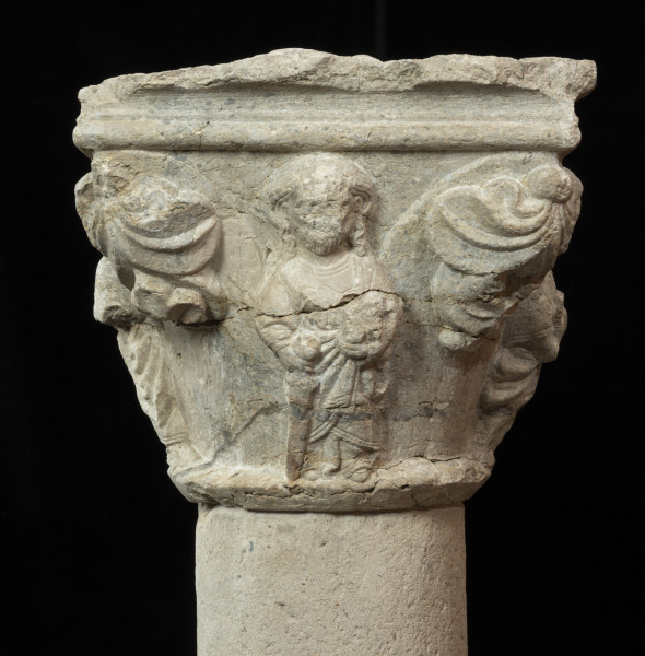 kapitel z przedstawieniem czterech apostołów - Ujęcie czwartego apostoła z uszkodzoną twarzą. Kapitel blokowo-kielichowy, rozszerzający się ku górze, wykuty z jednego kawałka kamienia. Okrągłą podstawę otacza wałek o profilu trójkątnym. Zwieńczenie w formie szerokiej czworobocznej płyty abakusa o krawędziach profilowanych wałkiem i wklęską. Na osi każdego boku półplastyczne przedstawienie frontalnie ukazanych postaci Apostołów: Piotra, Pawła, Bartłomieja i Jakuba Starszego. Pomiędzy nimi duże liście palmetowe dekoracyjnie ułożone na narożnikach. Postaci o dużych głowach i dość smukłej budowie, trzymają atrybuty. Długie tuniki i płaszcze o pofałdowanej powierzchni przylegają do sylwetek. Charakterystyczne zdobienie krawędzi szat podwójnym wałkiem i symetrycznie ułożone falujące fryzury. Relief półplastyczny, światłocieniowy. Tło opracowane gładko. Jedno z naroży kapitela uszkodzone. Liczne pęknięcia i ukruszenia na powierzchni kamienia. Pod spodem centralne wgłębienie do umocowania trzpienia łączącego kapitel z trzonem.