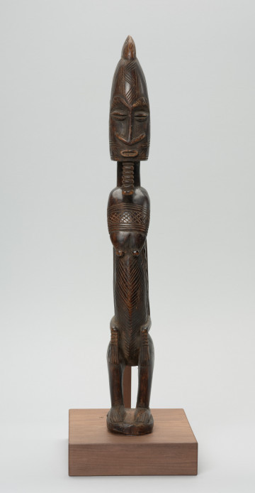 figura - postać przodka - Ujęcie z przodu. Drewniana rzeźba kobiety o zgeometryzowanej formie zdobiona licznymi grupami nacięć. Sylwetka spłaszczona po bokach.