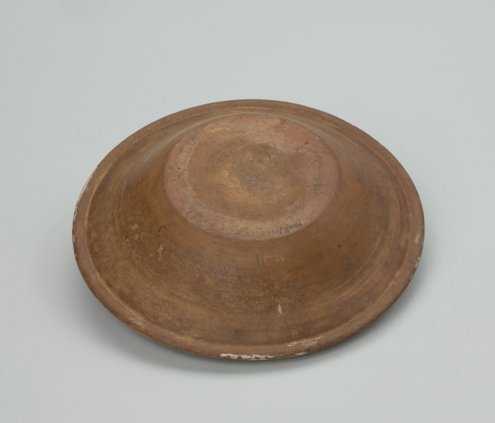 talerz - Ujęcie spodu  z góry. Talerz wykonany na kole garncarskim z gliny o jasnej barwie ( z małą zawartością tlenków żelaza). Dno niewyodrębnione przechodzi w lekko odchylony na zewnątrz korpus z wyraźnym kołnierzem. Szkliwiony tylko po wewnętrznej stronie i dekorowany odręcznie stylizowanym, jednoelementowym motywem roślinnym. W miejscu przejścia dna w korpus przypadkowa kropla farby. Wylew z kołnierzem zdobiony podwójną, miejscami niestaranna linią. Zdobienia w kolorach: dominującym niebieskim oraz zielonym.