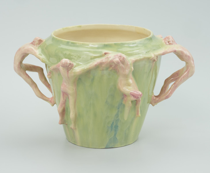 ceramika - Ujęcie z boku; Widoczne cztery postacie - figury ceramiczne przyczepione do malowanego w kolorze zielonym wazonie. Figura męska odwrócona plecami ciągnąca kobietę z prawej za włosy. Po bokach wazonu dwie męskie postacie ciągnące za ręke parę na przodzie wazonu.