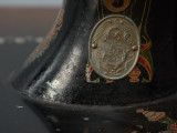 Maszyna do szycia ze stolikiem - Ujęcie z przodu, fragment górnej części; Maszyna do szycia - część szyjąca. Na nodze maszyny w metalowym owalu znajduje się emblemat Stoewera: litera S i B z malutkim smokiem w górnej części litery S, ozdobione ornamentem roślinnym.