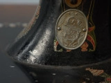 Maszyna do szycia ze stolikiem - Ujęcie z przodu, fragment górnej części; Maszyna do szycia - część szyjąca. Na nodze maszyny w metalowym owalu znajduje się emblemat Stoewera: litera S i B z malutkim smokiem w górnej części litery S, ozdobione ornamentem roślinnym.
