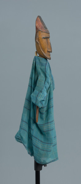 lalka teatralna: Donso (myśliwy) - Ujęcie z boku z prawej strony. Drewniana lalka przedstawiająca postać męską. Twarz podłużna, wąska, mocno zarysowane czarne brwi, długi nos, wąskie usta, niewielka broda i wąsy. Postać jest ubrana w długą niebieską szatę w kratę. Na głowie wysoka czapka. Dłonie z zaznaczonymi palcami.