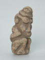 rzeźba; Figura kultu zmarłych - Ujęcie z przodu z prawej strony. Rzeźbiona w szarobeżowym steatycie siedząca postać ludzka z dużą głową o masywnych, okragłych okularach i przykurczonymi nogami, których kolana sięgają brody.