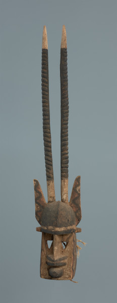 rzeźba , maska - Ujęcie z przodu. Część twarzowa: czoło wypukłe, czarne, cienki grzbiet nosa, szerokie nozdrza, stożkowate usta. wycięte trójkątne oczy. Na obu bokach części twarzowej wymalowany zyg-zakowaty wzór (czarna linia, wypełnienie trójkątów - czerwone). Część twarzowa zwieńczona lekko wygiętymi do tyłu długimi żłobkowanymi rogami oraz szpiczastymi uszami ozdobionymi zyg-zakowatymi wzorami (kolory analogicznie jak wyżej). W części tylnej maski wywiercone otwory, umożliwiające przymocowanie jej do twarzy tancerza oraz pozostałości po sznurku.