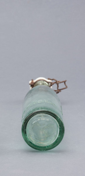 szklana butelka z kapslem - Ujęcie z dołu; Butelka piwówka z grubego szkła przeźroczystego półbiałego z odcieniem zielonym. Szyjka przechodząca łagodnie z korpusu zakończona przegrubieniem z wklęską na drut zamknięcia bocznego - dźwigniowego. Dno wklęsłe.