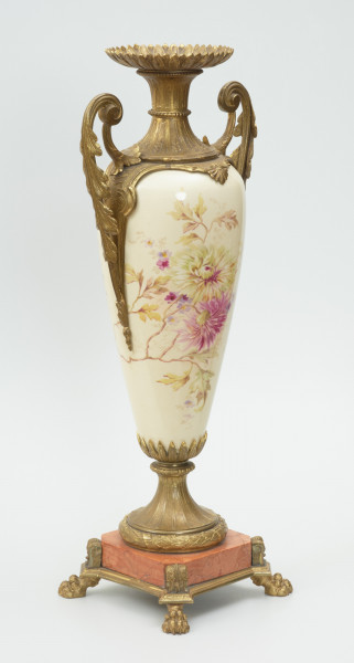 Waza - Ujęcie skosem w prawą stronę. Waza dekoracyjna o bogatej, barwnej dekoracji reliefowej, w kształcie kielicha ze spłaszczoną czarą z dwoma uchwytami i przykrywą. Podstawa okrągła, profilowana z czterema prostokątnymi poszerzeniami z listkiem akantu. Trzon na styku z podstawą ozdobiony wałkiem z liści akantu, w części środkowej wypełniony reliefem z dwiema brązowymi maskami i girlandami drobnych, barwnych kwiatów. W górnej części trzonu perełkowy otok z przewężeniem na styku z czarą. Czara z szerokim brzegiem ozdobionym dekoracją geometryczną i zwężającym się do dołu brzuścem dekorowanym dwiema wypełniającymi całą wysokość czary brązowymi maskami, których zielone grzywy w formie palmety opierają się na górnej krawędzi brzegu, a brody na spodzie czary ozdobionym granatowymi 