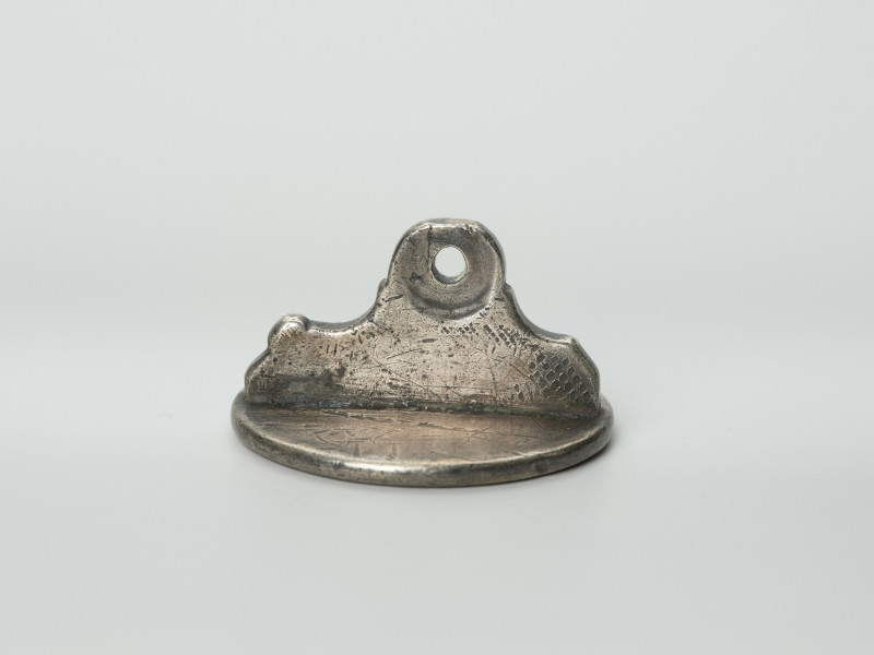 tłok pieczętny - Ujęcie z boku tyłu pieczęcią w dół. Okrągły tłok pieczętny ze srebra z uchwytem w formie profilowanej płytki oraz wizerunkiem łba gryfa i gotyckim napisem w polu pieczęci.