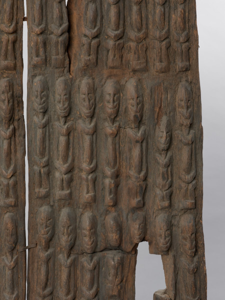 drzwi - Ujęcie z przodu z lewej strony w przybliżeniu. Drzwi drewniane do spichlerza, składające się z dwóch części połączonych klamerkami z żelaznej blachy. Zdobione płaskorzeźbionymi postaciami o schematycznych rysach z dłońmi na kolanach. Postaci w pięciu poziomych rzędach po 12 i 13 w rzędzie. Na wysokości czwartego rzędu przymocowana z lewej strony nieruchoma część zasuwy, wykonana z drewna i zdobiona rzeźbioną siedzącą postacią. Ruchomej części zasuwy brak.