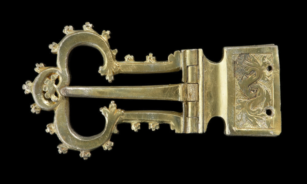 Pozłacana sprzączka dwuczęściowa na zawiasie z trójlistną ramą - Ujęcie z przodu. Pozłacana sprzączka dwuczęściowa na zawiasie z trójlistną ramą. Rama symetryczna, długa, trójlistna, z regularnie ułożonymi na zewnątrz wypustkami. Skuwka uformowana w prostokąt, na który naniesiono motyw liścia akantu. Tło pokryto skośną kratką.