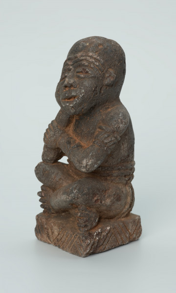 Ujęcie z przodu z lewej. Rzeźbiona w grafitowym kamieniu postać ludzka - najprawdopodobniej mężczyzna - w pozycji siedzącej z nogami skrzyżowanymi. Rzeźba ma charakterystyczne bransolety – dwie na ramionach oraz jedną na nadgarstku - które ozdobiono wzorem w postaci ukośnych linii. Widoczne ślady czerwonego piasku, rysy, mikropęknięcia oraz ubytek z tyłu głowy.