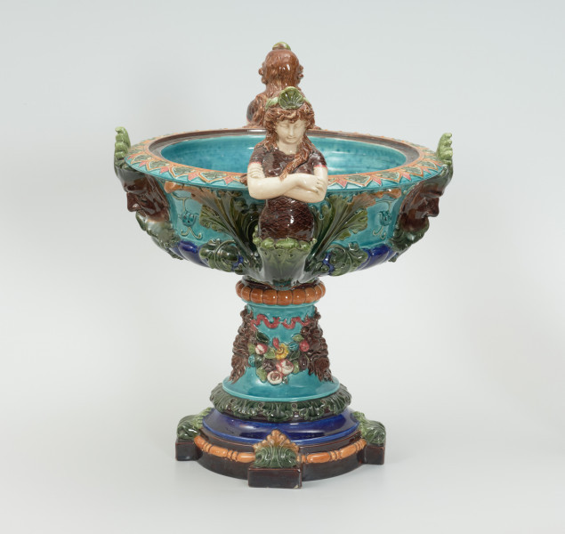 Waza - Ujęcie z lewej strony. Waza w kształcie smukłej amfory. Brzusiec ceramiczny, rozszerzający się ku górze, w kolorze kremowym, malowany podszkliwnie (z uzupełnieniami naszkliwnymi) we wzory kwiatowe - bukiety barwnych kwiatów w typie aster w kolorach fioletowo-czerwonym, liliowym, żółtym i białym, z gałązkami i listkami oraz drobnymi kwiatkami przypominającymi bratki o barwie fioletowo-czerwonej i liliowej. Górna część wazy wraz z uchwytami oraz stopą i część podstawy wykonane jako odlewy z mosiądzu. Mosiężna szyja wąska z otokiem perełkowym w środku ozdobiona jest pionowym, rozszerzającym się ku dołowi i ku górze rytmicznym podziałem z ornamentem geometryczno-roślinnym. Na pogrubionym skraju szyi umieszczona jest płytka miseczka w formie kwiatu o wąskich płatkach, stanowiąca wylewkę wazonu. Poniżej dolnej części szyi oprawa wierzchołka brzuśca składająca się z otoku o sercowym wycięciu zwieńczonym profilowaną palmetką oraz dwóch wydłużonych, sięgających 1/3 wysokości brzuśca, pionowych wsporników uchwytów. Te ostatnie składają się z 2 części: zewnętrzna wychodząca ze wsporników opartych na brzuścu, wyniesiona jest po niewielkim skosie do połowy wysokości szyi i na końcu wywinięta do wnętrza wolutowo, a druga - krótsza, skierowana ku dołowi łączy łagodnym łukiem wolutę z górną częścią oprawy brzuśca, kończąc się wywiniętymi na zewnątrz listkami. Uchwyty żłobkowane, zdobione motywem liści. Wazon wsparty jest na mosiężnej podstawie, składającej się z wieńca oplecionego wstęgą, na którym spoczywa zwężająca się ku górze stopa o brzegach ozdobionych perełkowaniem i pierścieniem, wspierającym koszyczek, ujmujący dolną część ceramicznego brzuśca, w formie kwiatu o ostro zakończonych, wąskich płatkach. Całość spoczywa na podstawie z kwadratowej płytki czerwonego marmuru ze ściętymi narożnikami, ujętymi mosiężnymi wspornikami ramy wspierającej płytę od dołu i opartej w narożach na czterech lwich łapach.