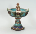 Waza - Ujęcie z lewej strony. Waza w kształcie smukłej amfory. Brzusiec ceramiczny, rozszerzający się ku górze, w kolorze kremowym, malowany podszkliwnie (z uzupełnieniami naszkliwnymi) we wzory kwiatowe - bukiety barwnych kwiatów w typie aster w kolorach fioletowo-czerwonym, liliowym, żółtym i białym, z gałązkami i listkami oraz drobnymi kwiatkami przypominającymi bratki o barwie fioletowo-czerwonej i liliowej. Górna część wazy wraz z uchwytami oraz stopą i część podstawy wykonane jako odlewy z mosiądzu. Mosiężna szyja wąska z otokiem perełkowym w środku ozdobiona jest pionowym, rozszerzającym się ku dołowi i ku górze rytmicznym podziałem z ornamentem geometryczno-roślinnym. Na pogrubionym skraju szyi umieszczona jest płytka miseczka w formie kwiatu o wąskich płatkach, stanowiąca wylewkę wazonu. Poniżej dolnej części szyi oprawa wierzchołka brzuśca składająca się z otoku o sercowym wycięciu zwieńczonym profilowaną palmetką oraz dwóch wydłużonych, sięgających 1/3 wysokości brzuśca, pionowych wsporników uchwytów. Te ostatnie składają się z 2 części: zewnętrzna wychodząca ze wsporników opartych na brzuścu, wyniesiona jest po niewielkim skosie do połowy wysokości szyi i na końcu wywinięta do wnętrza wolutowo, a druga - krótsza, skierowana ku dołowi łączy łagodnym łukiem wolutę z górną częścią oprawy brzuśca, kończąc się wywiniętymi na zewnątrz listkami. Uchwyty żłobkowane, zdobione motywem liści. Wazon wsparty jest na mosiężnej podstawie, składającej się z wieńca oplecionego wstęgą, na którym spoczywa zwężająca się ku górze stopa o brzegach ozdobionych perełkowaniem i pierścieniem, wspierającym koszyczek, ujmujący dolną część ceramicznego brzuśca, w formie kwiatu o ostro zakończonych, wąskich płatkach. Całość spoczywa na podstawie z kwadratowej płytki czerwonego marmuru ze ściętymi narożnikami, ujętymi mosiężnymi wspornikami ramy wspierającej płytę od dołu i opartej w narożach na czterech lwich łapach.