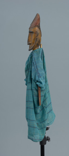 lalka teatralna: Donso (myśliwy) - Ujęcie z boku z lewej strony. Drewniana lalka przedstawiająca postać męską. Twarz podłużna, wąska, mocno zarysowane czarne brwi, długi nos, wąskie usta, niewielka broda i wąsy. Postać jest ubrana w długą niebieską szatę w kratę. Na głowie wysoka czapka. Dłonie z zaznaczonymi palcami.