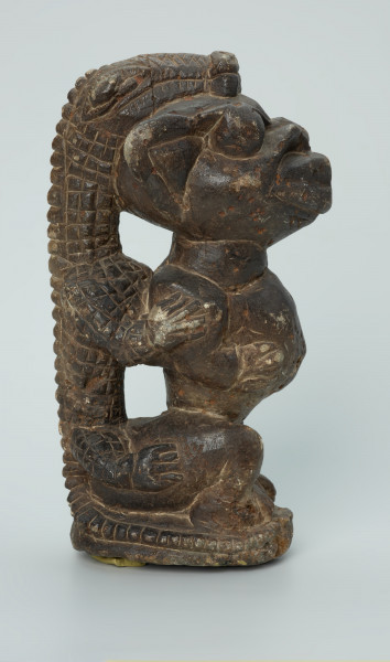 rzeźba; przedmiot obrzędowy; Figura kultu sił wegetacji - Ujęcie prawy bok. Rzeźbiona w grafitowym steatycie, siedząca na owalnej podstawie, postać ludzka, którą od tyłu obejmuje postać zoomorficzna o cechach jaszczurki.