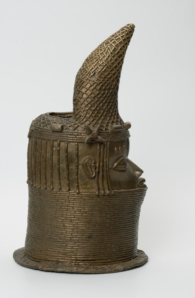 Głowa Królowej Matki - Ujęcie z boku z prawej strony. Brązowy, pusty w środku odlew głowy kobiety, o złotym zabarwieniu. Twarz o cechach negroidalnych, wydatnych, lekko rozchylonych ustach oraz szerokim u nasady nosie ma oczy z wyraźnie zaznaczoną linią powiek. Na ich wysokości usytuowane są nieduże uszy. Lekko wypukłe czoło dekorowane jest pionowymi inkrustacjami z żelaza oraz trzema podłużnymi, symetrycznie rozmieszczonymi, pogrubieniami nad każdym okiem. Szyję i podbródek do wysokości dolnej wargi zasłonięto masywnym naszyjnikiem z korali. Głowa jest nienaturalnie poszerzona w tylnej części, tuż pod przechyloną do przodu koroną o charakterystycznym kształcie dziobu papugi, którą nad uszami ozdobiono dwoma rozetami. W dolnej części korony widnieje pięć ozdób o walcowatej formie, trzy nad czołem, dwie za rozetami oraz opaska z korali. Tuż za koroną znajduje się duży, owalny otwór na rzeźbiony kieł słonia, którego brak. Poniżej, po obu stronach twarzy i z tyłu głowy, zaznaczono zaplecione w warkoczyki włosy. Z tyłu głowy pomiędzy włosami a naszyjnikiem usytuowany jest niewielki, okrągły otwór. U podstawy odlewu znajduje się pozioma listwa, dekorowana motywem plecionki.