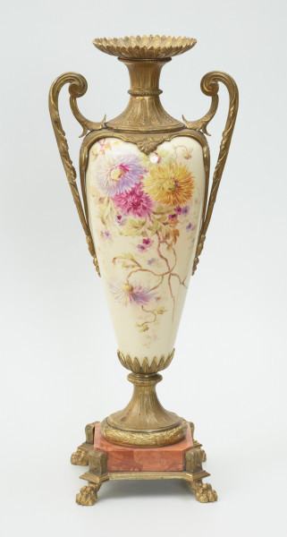 Waza - Ujęcie z przodu. Waza dekoracyjna o bogatej, barwnej dekoracji reliefowej, w kształcie kielicha ze spłaszczoną czarą z dwoma uchwytami i przykrywą. Podstawa okrągła, profilowana z czterema prostokątnymi poszerzeniami z listkiem akantu. Trzon na styku z podstawą ozdobiony wałkiem z liści akantu, w części środkowej wypełniony reliefem z dwiema brązowymi maskami i girlandami drobnych, barwnych kwiatów. W górnej części trzonu perełkowy otok z przewężeniem na styku z czarą. Czara z szerokim brzegiem ozdobionym dekoracją geometryczną i zwężającym się do dołu brzuścem dekorowanym dwiema wypełniającymi całą wysokość czary brązowymi maskami, których zielone grzywy w formie palmety opierają się na górnej krawędzi brzegu, a brody na spodzie czary ozdobionym granatowymi 