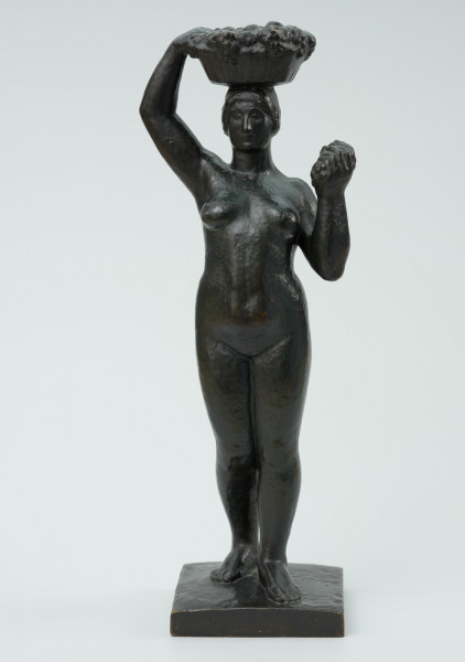 akt kobiecy - Ujęcie z przodu; Posążek przedstawia postać stojącej kobiety o pełnych kształtach dźwigającą kosz na głowie. Sylwetka wyprostowana o masywnych nogach, biodrach i niewielkich piersiach, zupełnie naga, z okrągłą twarzą i krótkich włosach- prawa noga lekko cofnięta, lewa ręka zgięta w łokciu dzierży na wysokości barku kiść owoców, prawa uniesiona ręka przytrzymuje na głowie płaski okrągły niski kosz wypełniony owocami. Figurka stoi na kwadratowej niskiej podstawce.