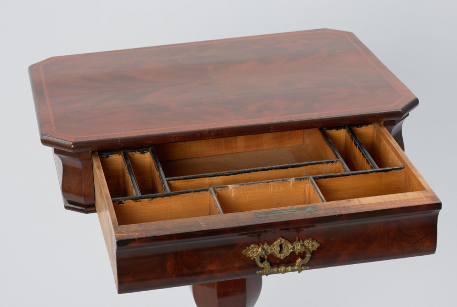 Stolik do robótek - Ujęcie blatu stolika z wysuniętą szufladką. Stolik z prostokątnym blatem ze ściętymi narożami i sfazowaną krawędzią, ozdobiony intarsją w formie wąskiego otoku z geometrycznym wzorem. Ścianka pod blatem o wybrzuszonej linii przekroju kryje szufladę z mosiężnym okuciem z dziurką do klucza i ruchomym uchwytem w centrum ścianki czołowej. Stolik wsparty na nodze w formie wydłużonej ośmiobocznej tralki gruszkowej, opartej przewężonym profilem na ośmiobocznym trzonie, do którego przymocowane są po przekątnej cztery nóżki składające się z dwóch odwróconych i połączonych esownic. Wewnątrz stolika znajduje się szuflada, która podzielona jest na jedenaście symetrycznie rozmieszczonych przegródek różnej wielkości, z krawędziami podmalowanymi na czarno.