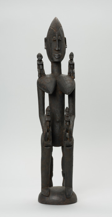 MNS/AF/2759 drewniana figura - Ujęcie z przodu. Drewniana, rzeźbiona figura kobiety. Na jej ramionach i rękach znajdują się mniejsze figurki.