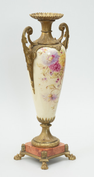 Waza - Ujęcie z tyłu skosem w lewo. Waza dekoracyjna o bogatej, barwnej dekoracji reliefowej, w kształcie kielicha ze spłaszczoną czarą z dwoma uchwytami i przykrywą. Podstawa okrągła, profilowana z czterema prostokątnymi poszerzeniami z listkiem akantu. Trzon na styku z podstawą ozdobiony wałkiem z liści akantu, w części środkowej wypełniony reliefem z dwiema brązowymi maskami i girlandami drobnych, barwnych kwiatów. W górnej części trzonu perełkowy otok z przewężeniem na styku z czarą. Czara z szerokim brzegiem ozdobionym dekoracją geometryczną i zwężającym się do dołu brzuścem dekorowanym dwiema wypełniającymi całą wysokość czary brązowymi maskami, których zielone grzywy w formie palmety opierają się na górnej krawędzi brzegu, a brody na spodzie czary ozdobionym granatowymi 