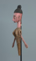 lalka teatralna: Yayoroba - Ujęcie z boku z lewej strony. Drewniana lalka przedstawiająca postać kobiecą. Różowa karnacja, mocno zaznaczone brwi, powieki górne, białe oczy z czarnymi źrenicami. Czarne, wysoko upięte włosy z pięcioma wypustkami. Odstające uszy, długi nos i wąskie usta. Zaznaczona cienka czarna broda. Z uszu zwisają długie kolczyki z kolorowych koralików. Szyja jest  nieproporcjonalnie długa. Lalka ma na sobie brązową sukienkę.