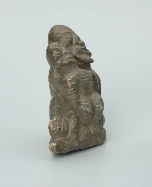 figura kultu przodków - Ujęcie z tyłu. Rzeźbione w grafitowym kamieniu dwie postaci ludzkie w pozycji siedzącej. Mniejsza postać – najprawdopodobniej dziecko - siedzi na kolanach większej - najprawdopodobniej kobiety. Obie postacie ozdobiono licznymi nacięciami liniowymi. Widoczne rysy i mikropęknięcia.
