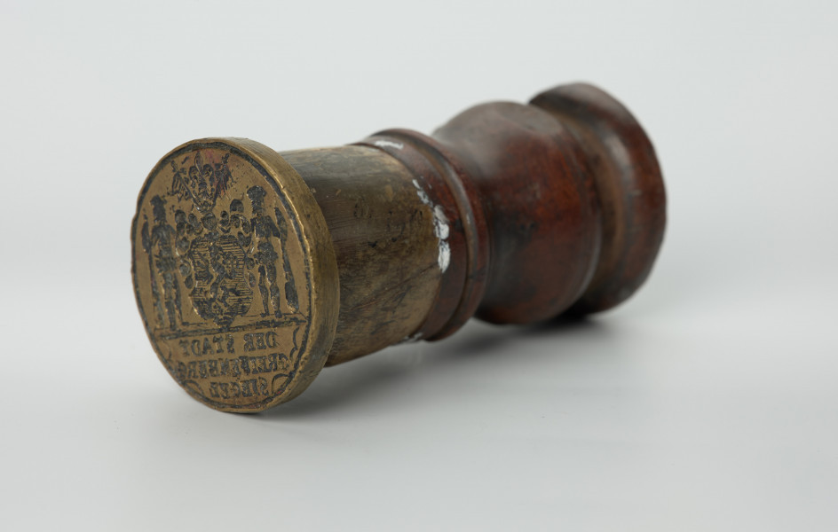 tłok pieczętny - Ujęcie z przodu skosem w lewą stronę. Okragły tłok pieczętny z brązu z drewniano-rogowym uchwytem oraz rysunkiem herbu Białogardu trzymanym przez dzikich ludzi w polu pieczęci.