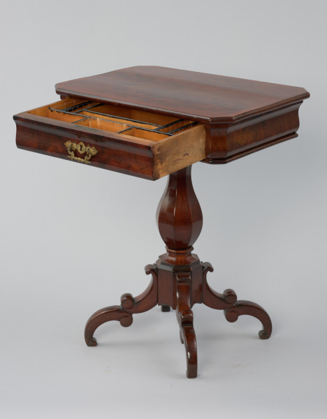 Stolik do robótek - Ujęcie z przodu skosem w lewo lekko z góry z wysuniętą szufladką. Stolik z prostokątnym blatem ze ściętymi narożami i sfazowaną krawędzią, ozdobiony intarsją w formie wąskiego otoku z geometrycznym wzorem. Ścianka pod blatem o wybrzuszonej linii przekroju kryje szufladę z mosiężnym okuciem z dziurką do klucza i ruchomym uchwytem w centrum ścianki czołowej. Stolik wsparty na nodze w formie wydłużonej ośmiobocznej tralki gruszkowej, opartej przewężonym profilem na ośmiobocznym trzonie, do którego przymocowane są po przekątnej cztery nóżki składające się z dwóch odwróconych i połączonych esownic. Wewnątrz stolika znajduje się szuflada, która podzielona jest na jedenaście symetrycznie rozmieszczonych przegródek różnej wielkości, z krawędziami podmalowanymi na czarno.