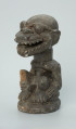 rzeźba; przedmiot obrzędowy; Figura kultu zmarłych - Ujęcie z przodu z lewej strony. Rzeźbiona w grafitowym steatycie, usadowiona na płaskiej, owalnej podstawie postać siedzącego z podkurczonymi nogami mężczyzny o masywnej głowie i tęgiej szyi z odsłoniętymi genitaliami. Powierzchnia rzeźby niejednolita; ubytek na prawym kolanie; ślady klejenia na twarzy.