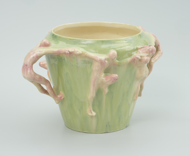 ceramika - Ujęcie z boku; Widoczna na wprost postać męska spoglądająca w lewą stronę, trzymająca za ręce postacie po bokach. Postać ma podciągnięte w górę lewe kolano. Postacie w kremowo-czeronym kolorze na tle zielonego wazonu.